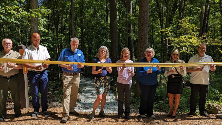 8 people, 3 men, 4 women, and another man, hold a ribbon and scissors. They are standing in front of a stand of trees on a mulched path with a new gravel path that leads to the new campsite behind them.