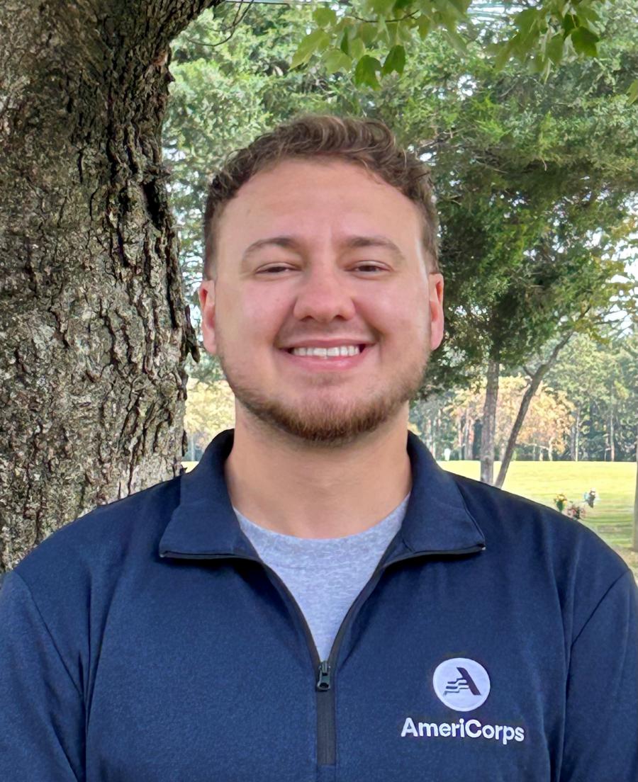 A white male in his 20s with wavy short hair and facial hair along the jaw line and upper lip. He is wearing a blue Americorp zip shirt.