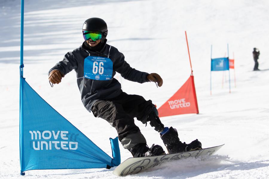 An adaptive snowboarder going down a mountain around some panels with the Move United logo