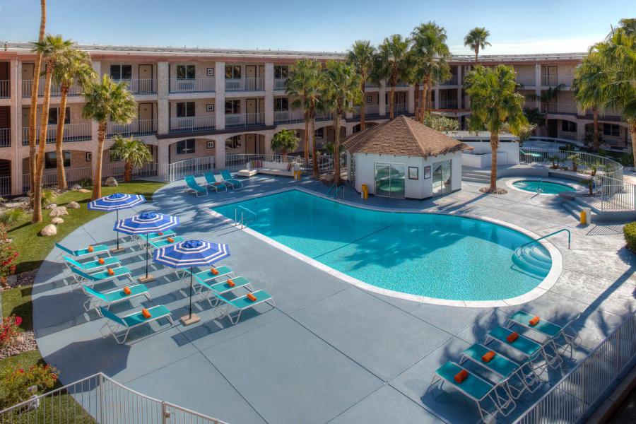 A hotel swimming pool with surrounding sun loungers and umbrellas, flanked by a three-story building with palm trees under a clear blue sky.