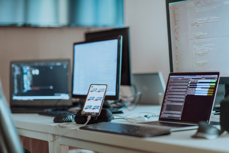 View of several computer screens and smartphone on a table with running programming code in an empty office.