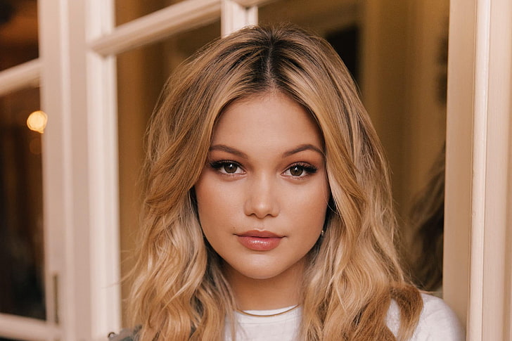 Close-up of Olivia Holt with long blonde hair styled in loose waves, wearing a neutral expression and light makeup, standing in front of a window with a warm indoor setting.