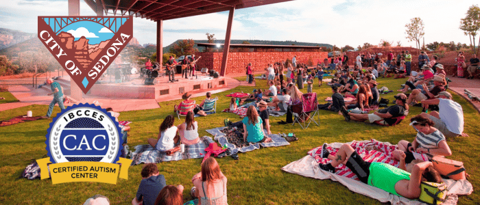 People of various ages sitting on the grass, engaged in a concert happening on a stage in the background.