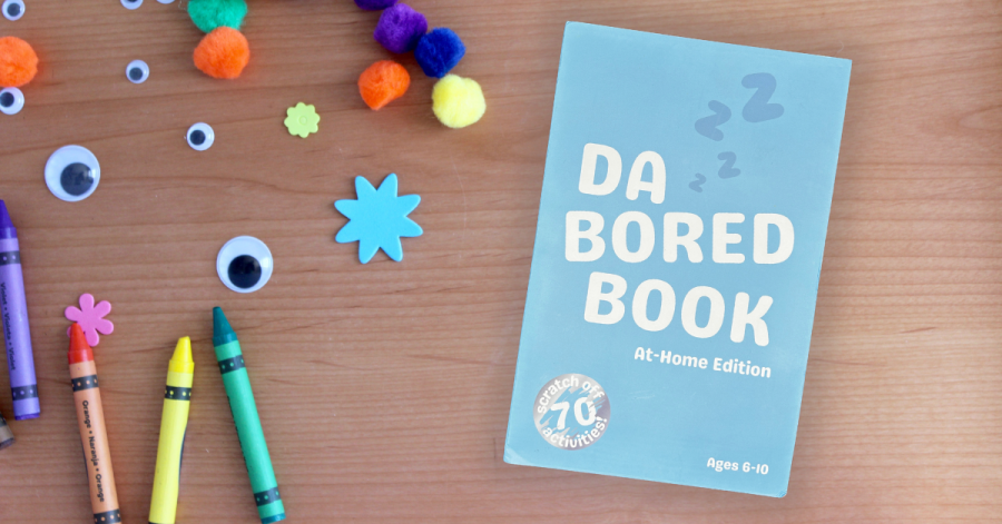 A copy of Da Bored Book: At-Home Edition placed on a table surrounded by craft supplies like crayons, googly eyes, and pom-poms. The book cover highlights "70 scratch-off activities" and is designed for children ages 6-10.