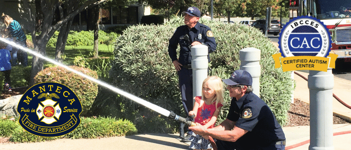 A firefighter from the Manteca Fire Department helps a young girl spray water from a fire hose, while another firefighter watches nearby, with the Manteca Fire Department and IBCCES Certified Autism Center™ logos displayed in the corners.