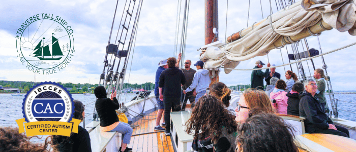 A group of people enjoy a scenic boat ride on a tall ship with the Traverse Tall Ship Co. and IBCCES Certified Autism Center™ logos displayed.