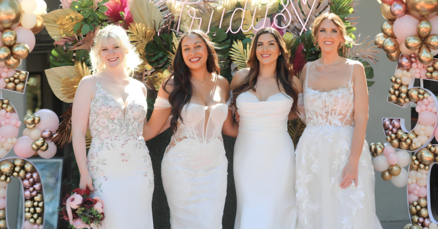 Four women wearing wedding dresses in front of a green backdrop that has the words "Trudys" on it. The photo is from Trudys Brides and Special Occasions 50th Anniversary Celebration.