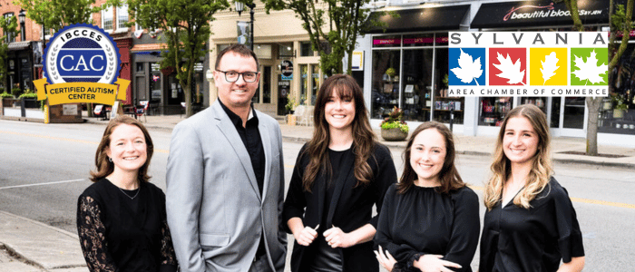 A group of professionals in black attire pose on a street, with local business signage and a chamber of commerce logo in the background.