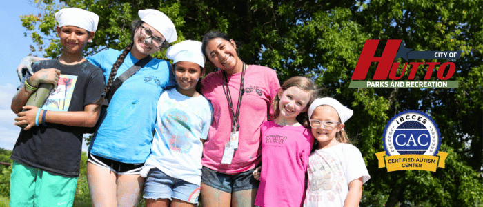 A group of children and adults in colorful shirts and hats pose outdoors, surrounded by greenery, promoting Hutto Parks and Recreation.