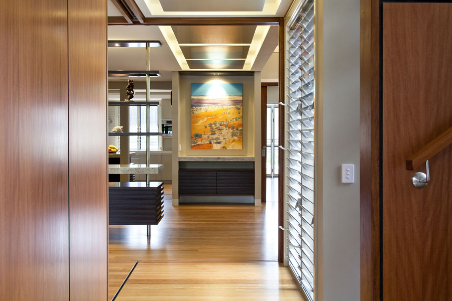 View through timber-lined entry to a modern interior with open shelving, louvre windows, feature artwork, and layered ceiling lighting.