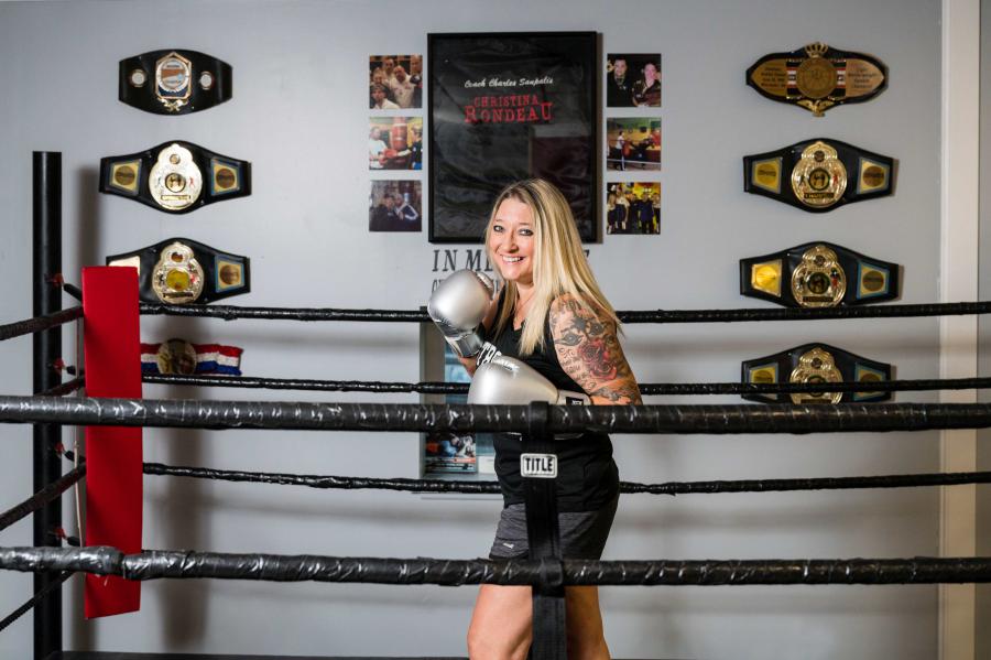 Professional Kickboxer and Fitness Trainer Christina Rondeau standing in a boxing ring with boxing gloves on and her Champion Belts are on a wall behind the ring