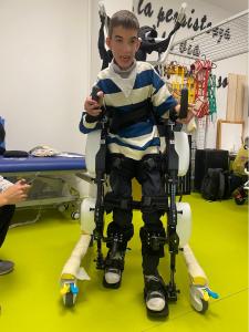 Child wearing RoboCT KidGo paediatric lower-limb exoskeleton stands upright without therapist nearby during supervised gait training at a Pisa rehabilitation centre.