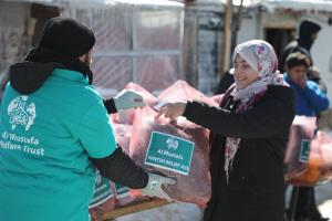A volunteer wearing a teal “Al Mustafa Welfare Trust” jacket gives a large pink winter aid package to a smiling woman in a headscarf, with snow-covered shelters in the background.