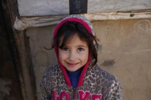 Close-up of a young girl with brown hair, wearing a leopard-patterned jacket and pink hood, smiling gently in front of a fabric-covered wall.