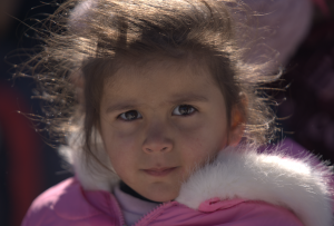 A close-up portrait of a young girl wearing a fluffy pink winter jacket, looking directly at the camera with windblown hair and a thoughtful expression.