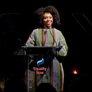 Chimamanda Ngozi Adichie smiling on stage behind a podium with the Equality Now logo on the front.