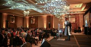 A man in a black-and-white patterned garment speaks at a clear podium during a formal gala. The ballroom glows with warm amber lighting. Guests in evening wear sit at round tables facing the stage, and a side screen shows his live image.