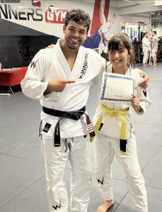 Young Brazilian Jiu-Jitsu student Jade Ghezal holding a belt promotion certificate with her coach at Winners Jiu-Jitsu Academy in Sunny Isles Beach, Florida. Both are wearing Winners-branded gis on the academy training mat.