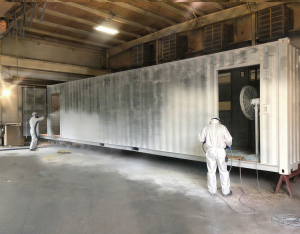 ArmorThane-certified applicators in full PPE spray-applying polyurea coating to a large steel enclosure inside a facility — authentic jobsite application of ArmorThane blast mitigation systems