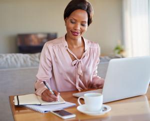 A woman wearing a light pink blouse writes in a notebook at her desk while working from home