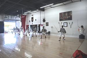 Students line up in fencing gear with masks during a saber class. 