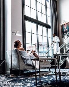Chris and Reg are both enjoying morning coffee in a beautiful loft style hotel suite that that has 20feet tall floor to ceiling windows. Chris is sitting in a chaise lounge wearing a white robe off off the right shoulder.  Reg is also wearing a white rob