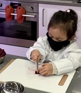 Image of a Little Kitchen Academy student (young Asian girl age 4) wearing a white chef coat cutting with the Little Kitchen Academy Child Safe Knife made of wood