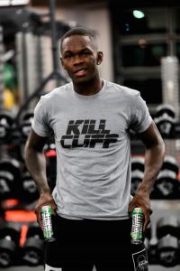 Israel Adesanya in a Kill Cliff shirt with Kill cliff beverage cans in a gym