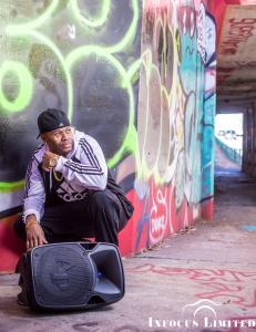 Artist Frequent-C is dressed in a black white Adidas track suit and matching sneakers while squatting near a large speaker in front of brightly-colored graffiti street art in Atlanta.
