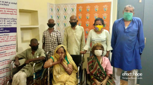 Photo: Two rows of people in India at a vaccine site. Back row from left to right: 2 men wearing black covid masks, a woman wearing a yellow covid mask and a man wearing a green mask. Front row: 3 individuals in wheelchairs. From left to right: one man we