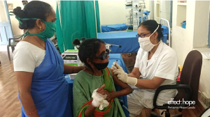 A nurse wearing white and a white covid-19 cloth mask inserts a needle into a woman wearing a green sari's arm. The woman receiving the vaccine is affected by leprosy. She wears dark glasses to assist her vision and has a bandage on one hand. A helper (wo