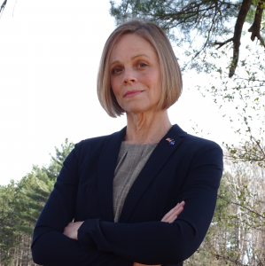 Headshot of Dr. Gillian Battino, a white woman with short blonde hair, wearing a black blazer and American flag pin standing with her arms crossed