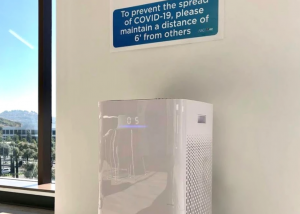 An image a large air purifier against an office wall, with a sign about preventing the spread of COVID-19 above it.