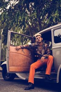 Marshall Hugh sitting in a vintage car facing towards the camera, wearing black boots, orange pants, a 70's style silk shirt, and gold chains.