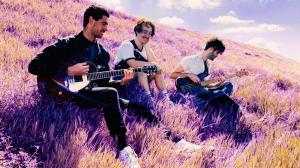 The three band members sat with guitars in hand in a meadow on a hill.