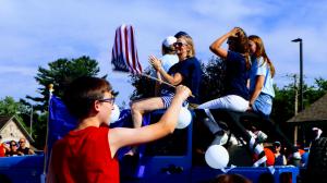 Dr. Battino her family and campaign staff sit atop a blue Jeep Wrangler in a parade precession as it passes by a young boy waiving an American Flag in the foreground