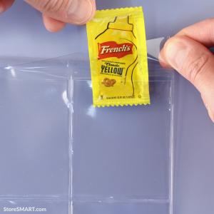 A protective flap is being lifted to insert a mustard packet into a clear plastic binder page.