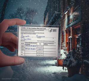 A closeup on a hand holding a vaccine card in a clear plastic protective sleeve. In the background, it's nighttime on a cozy, snowy city sidewalk.