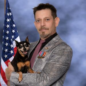 Photo of Mark Napier with American flag in the background holding his dog Sheila--a Miniature Pinscher. Napier is wearing his four top US Army medals, a CIA lapel pin and U.S. - Afghan flag lapel pin.