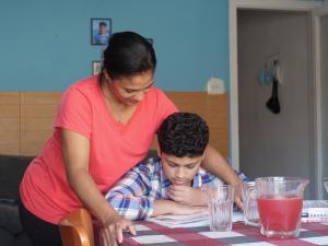 A mother is leaning over her son who is doing his homework in their colorful kitchen