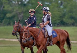 two women, one is a polo player and one is a polo umpire, on horses smiling and talking during a polo game