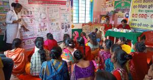 Group of women sitting on the floor in front of a woman who is teaching about menstrual health and hygiene, Camp Nydhee, India, April 19, 2022.