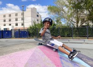 A boy gives the peace sign as he sits on top of his skateboard that will ride down a small hill.
