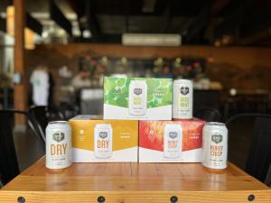 Photo of new package designs for Seattle Cider Co Dry, Basil Mint, and Honeycrisp in 6 pack boxes stacked with 12oz cans. Packaged cider is displayed on a table in the tasting room.