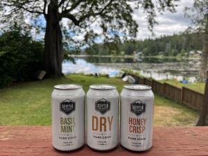 Three 12 oz cans from Seattle Cider, labeled Dry, Honeycrisp, and Basil Mint, sitting on a wooden fence overlooking a green yard with trees and a lake.