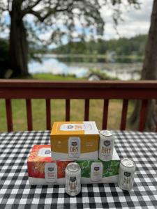Photo of Seattle Cider Co 12 oz cans and 6 pack boxes for Dry, Honeycrisp, and Basil Mint on a picnic table in a backyard overlooking trees and a lake