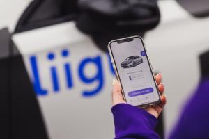 Woman holding a phone and showing Liigu contactless car rental app with a blurred car with Liigu logo in the background.  The app will open on the pickup screen where the user will see their assigned car and a button "I found the car" underneath.  th