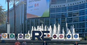 An image of the welcome flags and signs outside the entrance of the RE+ solar power conference in Anaheim, CA. There is a water fountain, palm trees, and a convention center building in the background. A few conference attendees sit on steps in the foreground.
