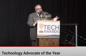 McQuillen wearing a gray top-hatted suit speaking into a microphone, the lectern has a sign that reads "TECH AWARDS" on the front page