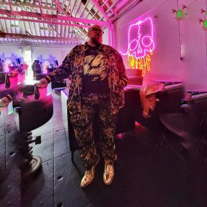 McQuillen stands in front of a pink neon skull display inside an art gallery, wearing a black and gold embellished tracksuit and metallic gold wingtip shoes